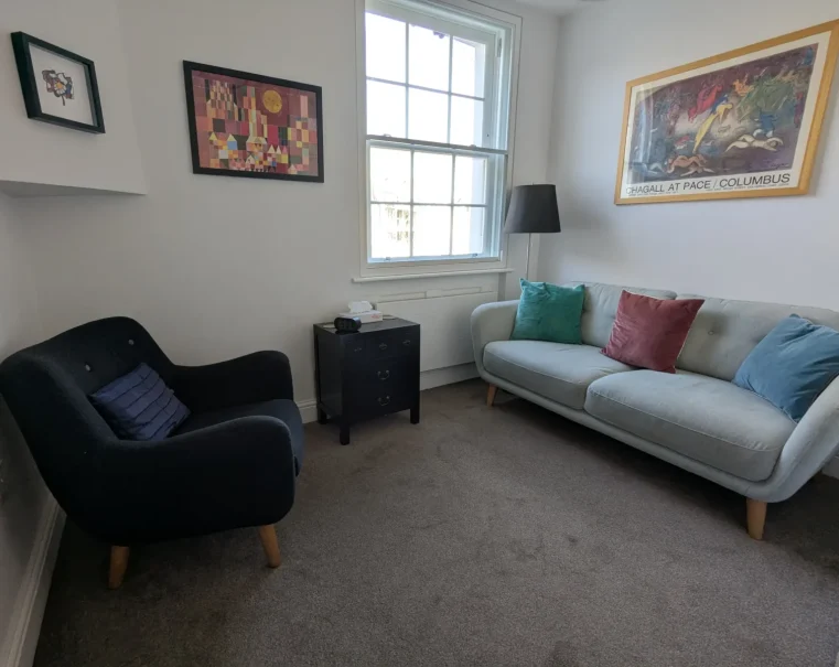 Bright counselling room with a grey armchair, a light grey sofa with colourful cushions, a small black side table, and framed artwork on white walls.